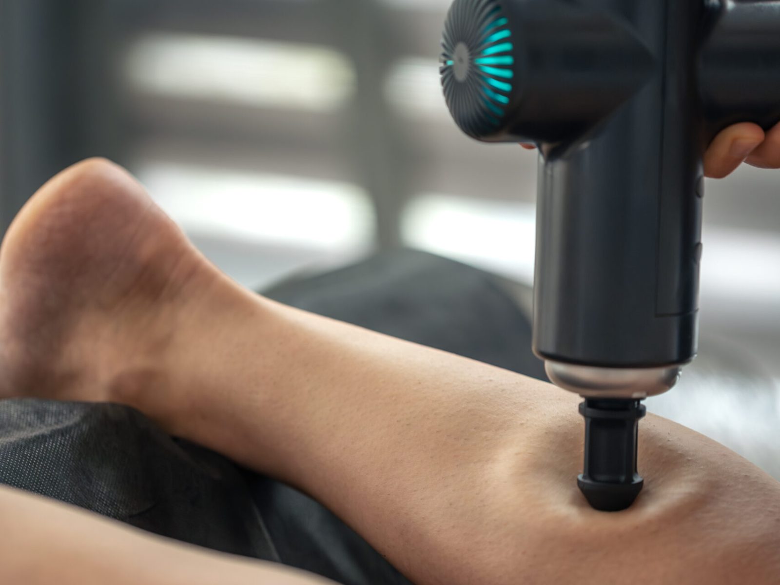 Close up of a percussion gun with cone head working the calf. Focused pressure for relaxation and mobility in a modern everyday self care setting.